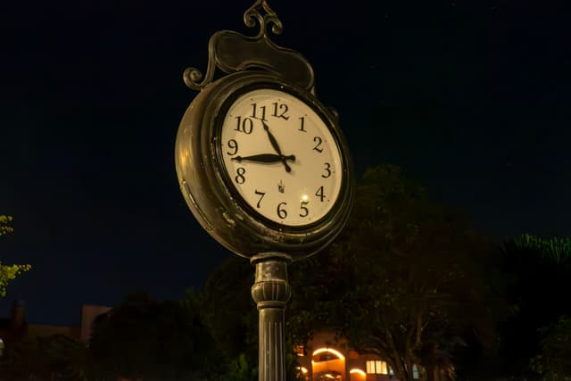 A vintage street clock displays the time as 8:20 against a dark night sky, with trees and softly lit buildings in the background