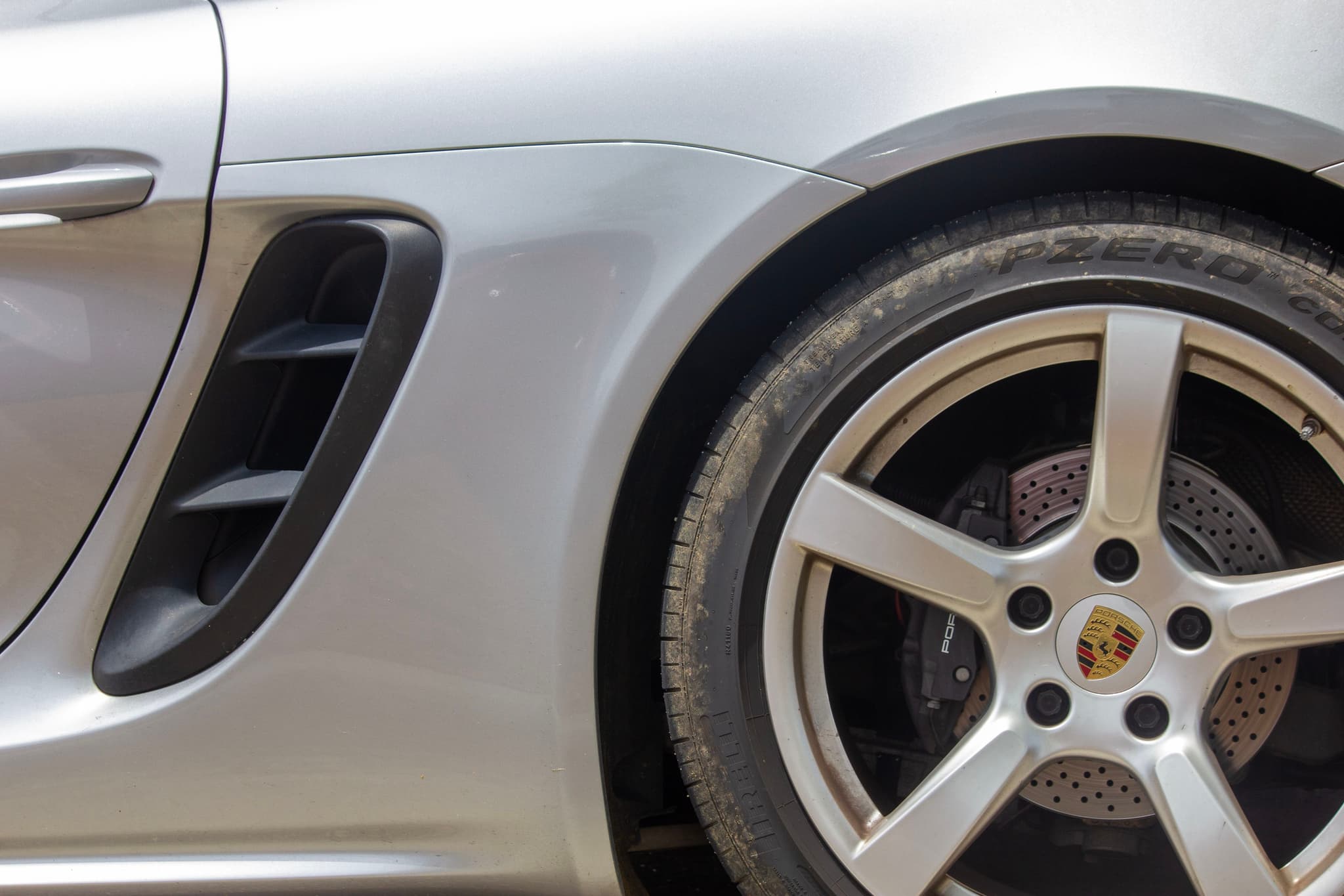 A close-up of a silver sports car's rear wheel and side vent, featuring a five-spoke alloy rim with a visible brake disc