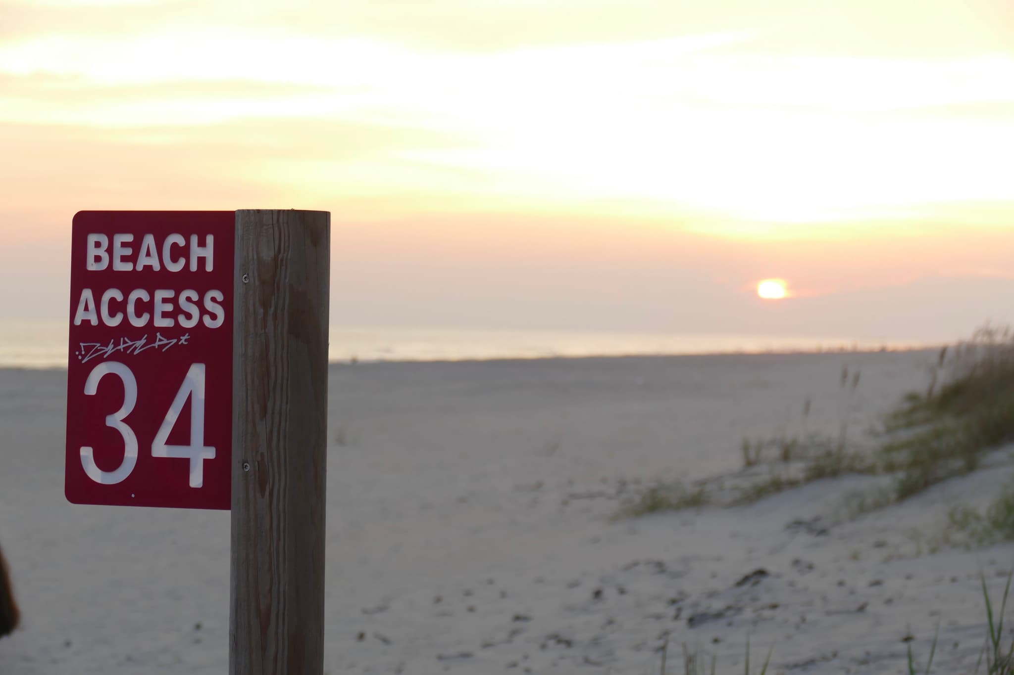 A beach access sign with the number 34 stands in the foreground, while the sun sets over the ocean in the background