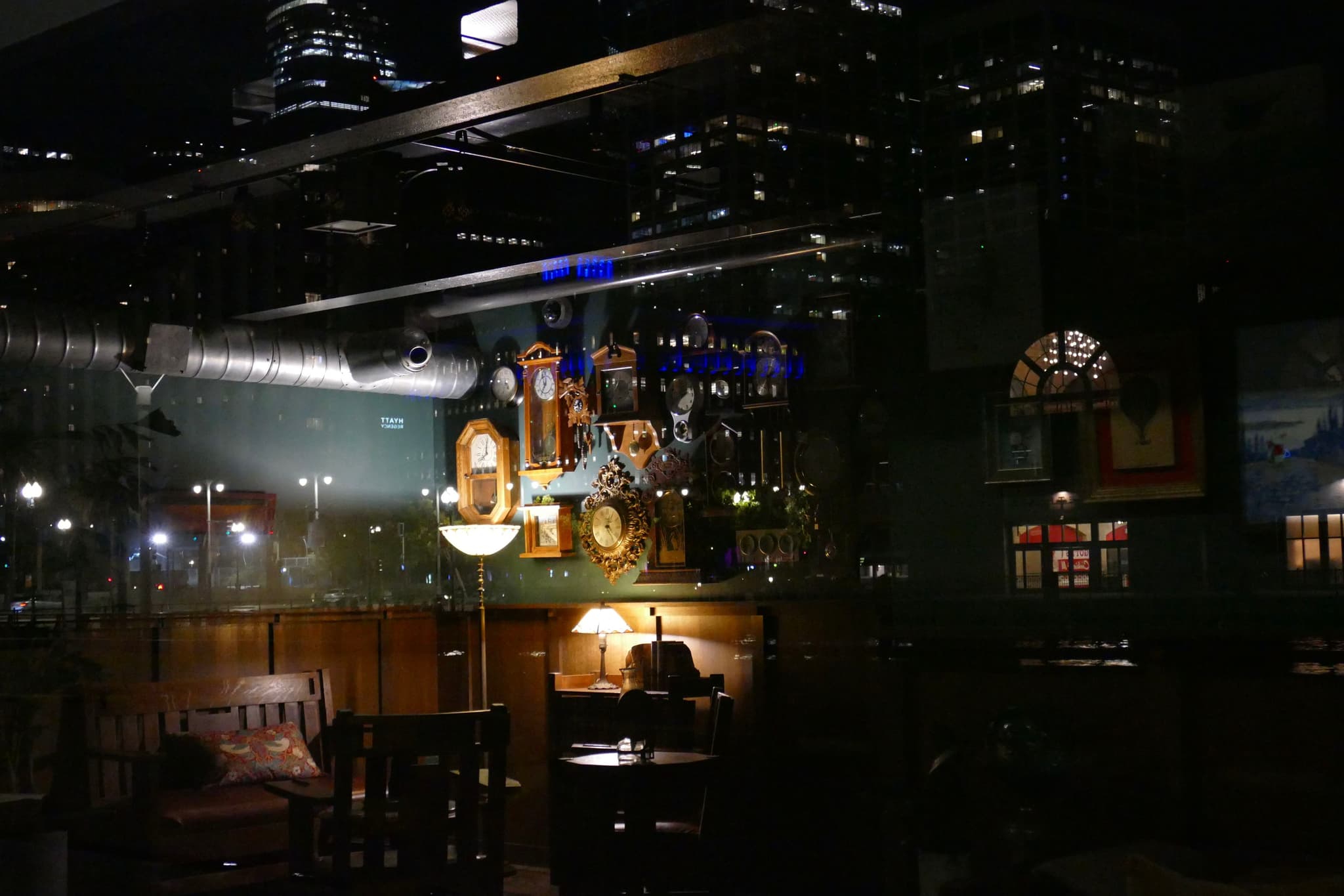 A dimly lit interior with wooden furniture and lamps, reflecting city lights and buildings in the glass