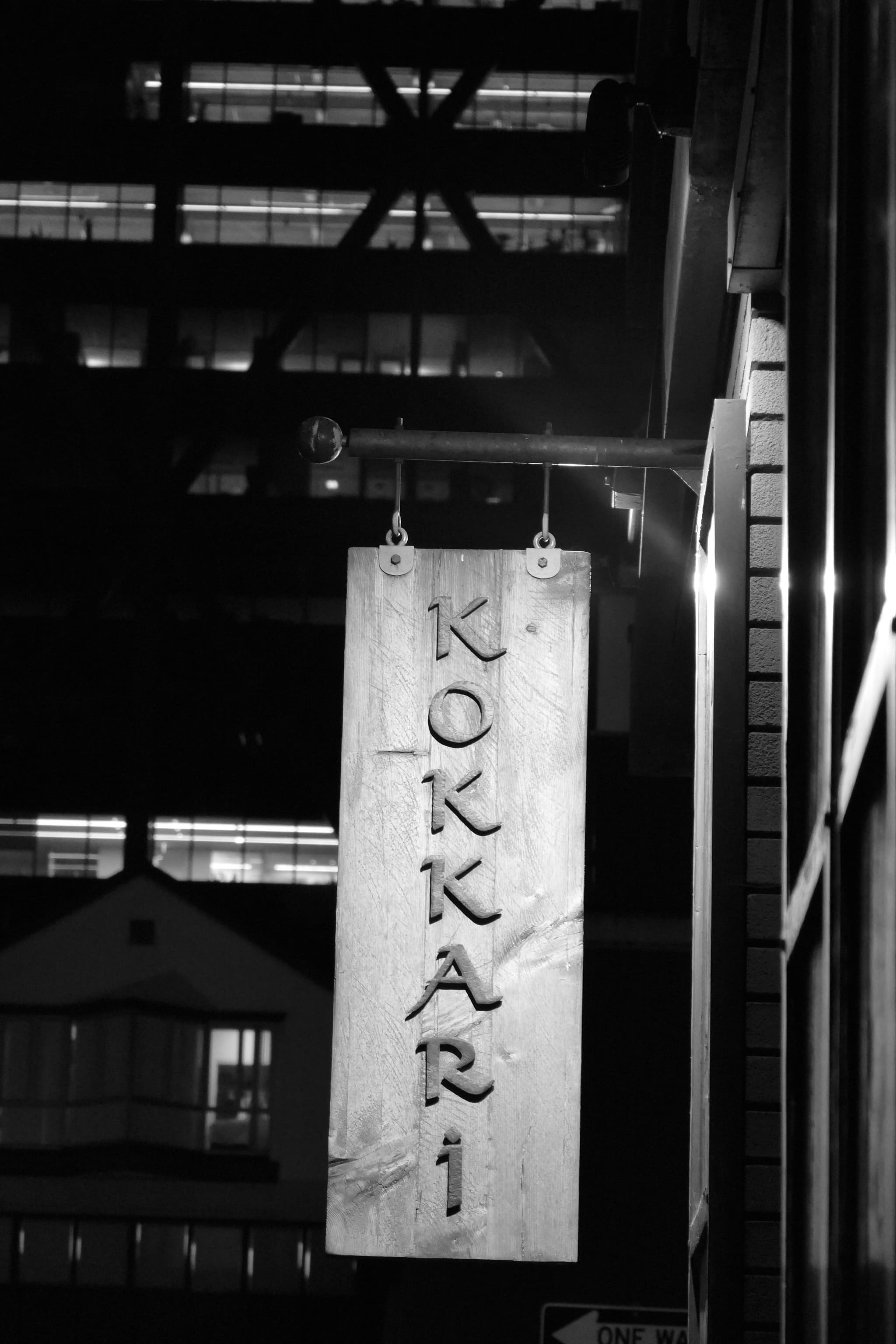 A vertical wooden sign with the word KOKKARI hangs on a building exterior, illuminated at night