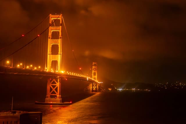 A brightly illuminated suspension bridge at night, with a dramatic, cloudy sky and reflections on the water below