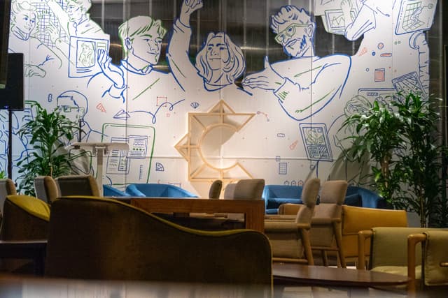 A modern lounge area with comfortable seating, plants, and a large mural featuring abstract figures on the wall